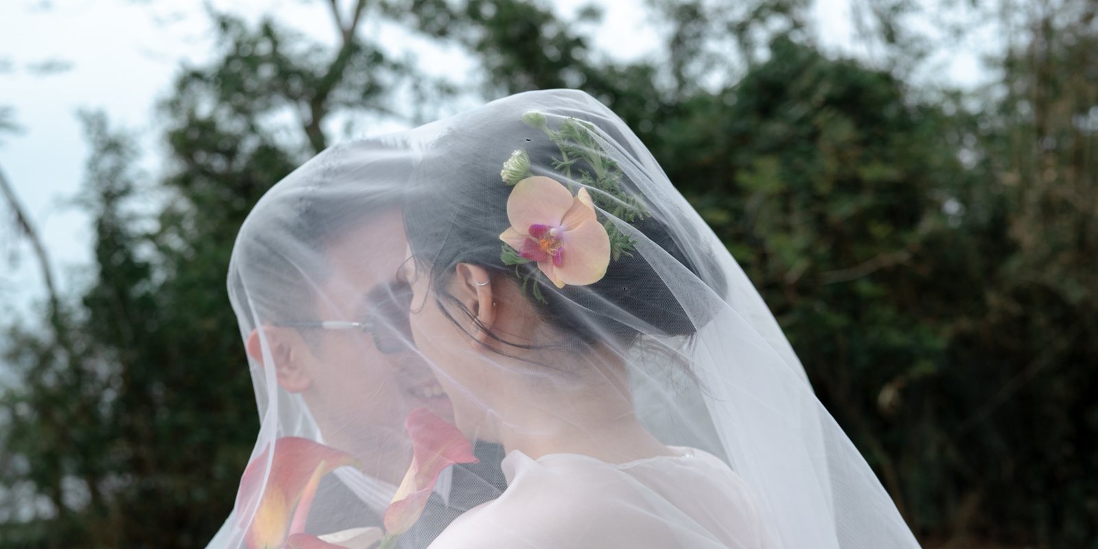 Bride and groom sharing a kiss under her veil outdoors, one year before their maternity session