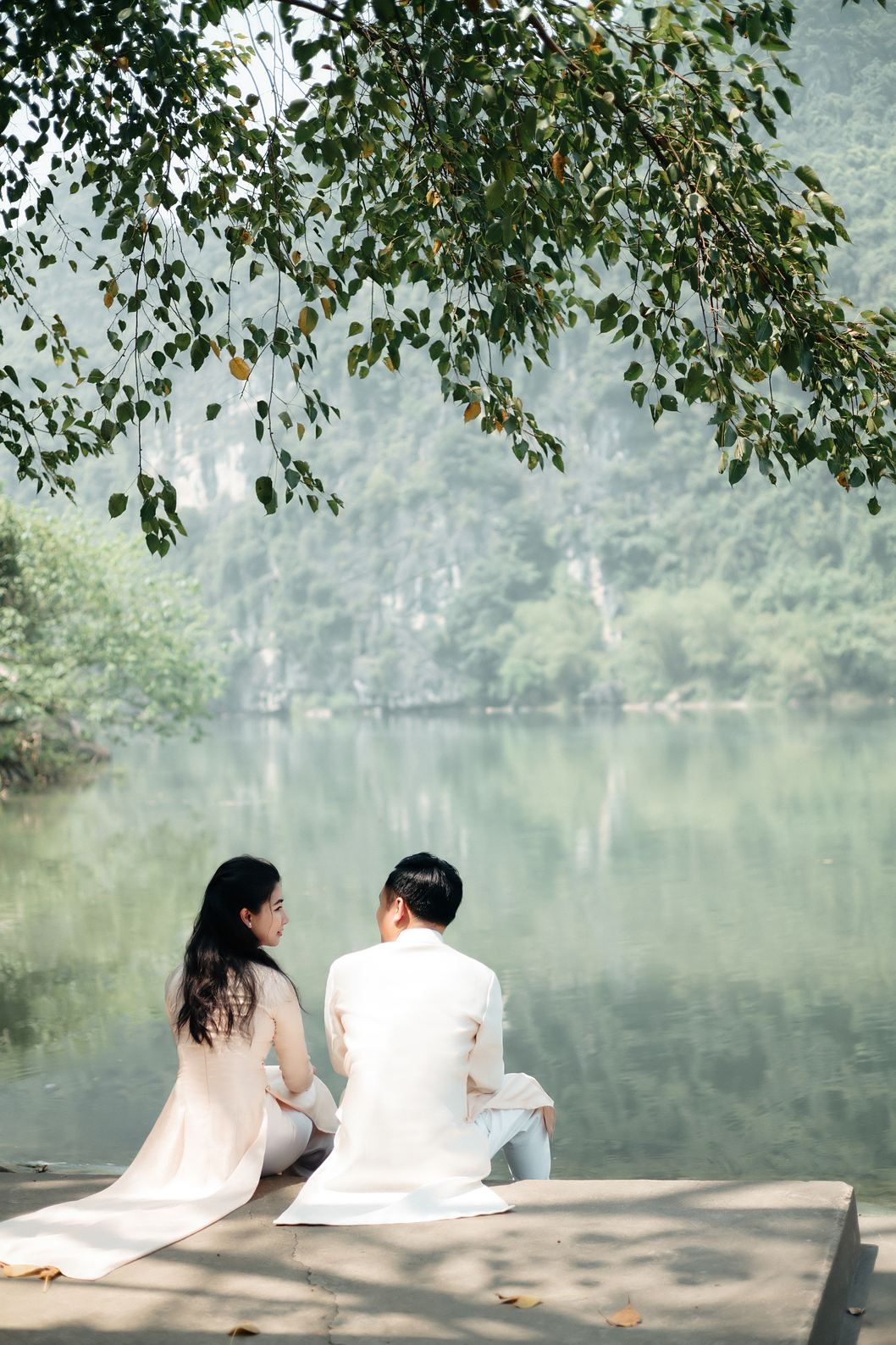 Cặp đôi áo trắng ngồi trên bến đá nhìn ra hồ và núi Ninh Bình