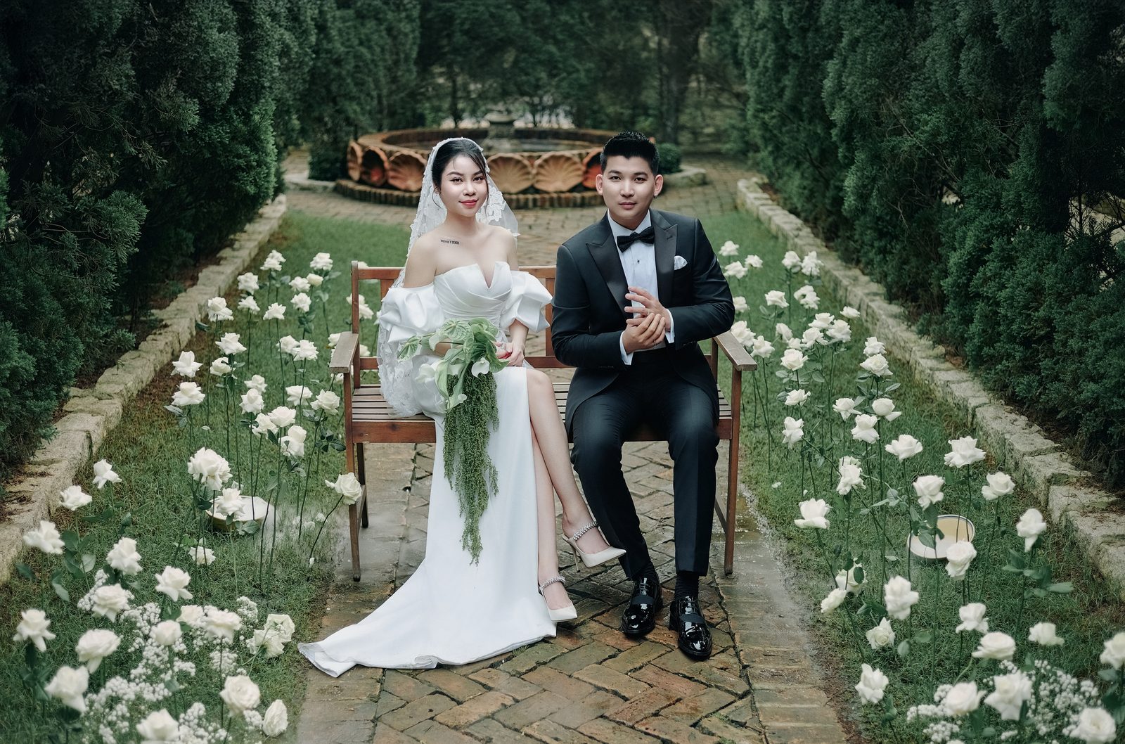 On a bench by the fountain, between beds of white roses