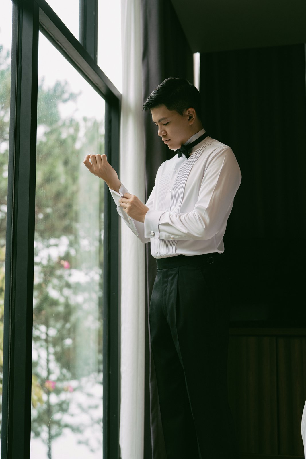 Groom fastening his cufflinks by the window while getting ready, documentary wedding Vietnam